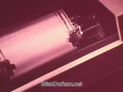 The still from 'Ohio Job Service PSA 3 - 1970's' depicts a close-up view of a mechanical device, likely a typewriter or printing machine, in operation. The focus is on the moving components, emphasizing the mechanical process of producing printed material. The color tone suggests a vintage aesthetic typical of the 1970s. The image conveys a sense of industry and productivity relevant to the context of job services.