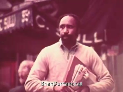 The still features a man walking in an outdoor setting, carrying a stack of papers and folders. He appears focused and engaged, wearing a light sweater. The background suggests an urban environment, possibly with storefronts visible. The overall tone of the image is warm and slightly nostalgic, reflective of the 1970s.