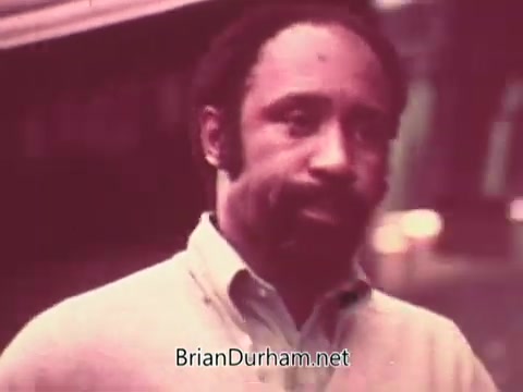 The still from 'Ohio Job Service PSA 3 - 1970s' features a man with a mustache and short-cropped hair, dressed in a light-colored sweater. He appears to be engaged in conversation, with a serious expression on his face. The image has a vintage quality, typical of 1970s film, with soft colors and graininess. The background is somewhat blurred, focusing attention on the individual.