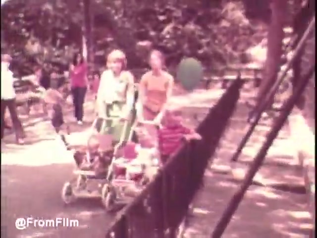 The still depicts a lively scene in a park from the early 1970s, showcasing children and adults enjoying the outdoors. Two young mothers are seen pushing double strollers, with babies inside. The background features other children playing, with one child grasping a balloon, adding to the cheerful atmosphere. The color palette reflects the warm, vibrant tones typical of 16mm film from that era. This captures the essence of family life and leisure, characteristic of the Pamper's Diapers brand messaging during that time.