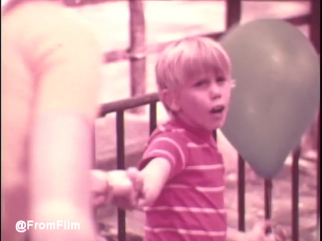 The still captures a moment from a 1970s Pamper's Diapers commercial, showcasing a young boy with blond hair wearing a red and white striped shirt. He appears to be excited or engaged as he reaches out with one hand, possibly holding onto a toy or a snack, while a colorful balloon floats nearby. The warm, soft color tones typical of 16mm film enhance the nostalgic feel of the scene, embodying the playful innocence of childhood.