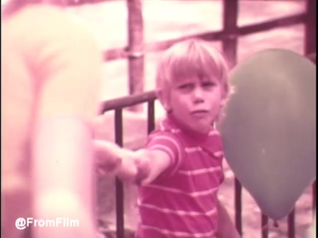 The still from the early 1970s 'Pamper's Diapers' commercial features a young boy in a striped shirt reaching out toward someone, possibly a caregiver or parent, while holding a balloon. The color palette appears warm and nostalgic, typical of that era's film style. The background suggests a playful outdoor setting, likely indicative of a family or picnic scenario, enhancing the theme of childhood and care associated with the product.