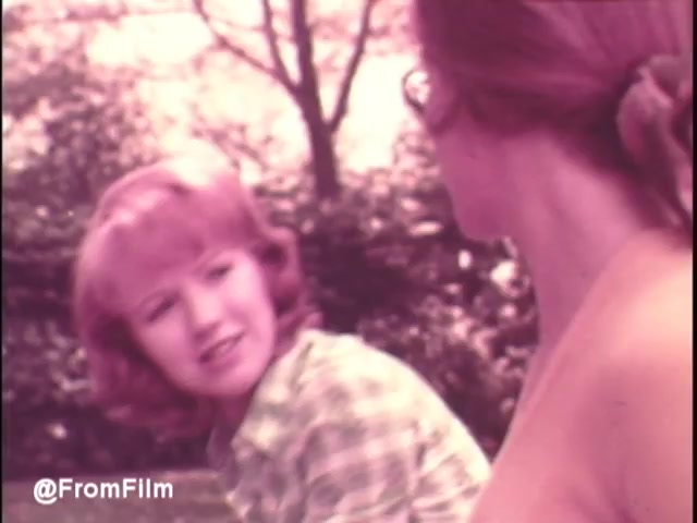 The still from the 'Pamper's Diapers commercial - early 1970s' depicts two women engaged in a light conversation outdoors. One has a playful expression, wearing a checkered shirt, while the other is partially visible and appears to be listening intently. The color palette has a vintage feel, typical of the 16mm film style, with soft hues that evoke a nostalgic atmosphere. The setting seems to be a park or a natural area, suggesting a familial or friendly context.