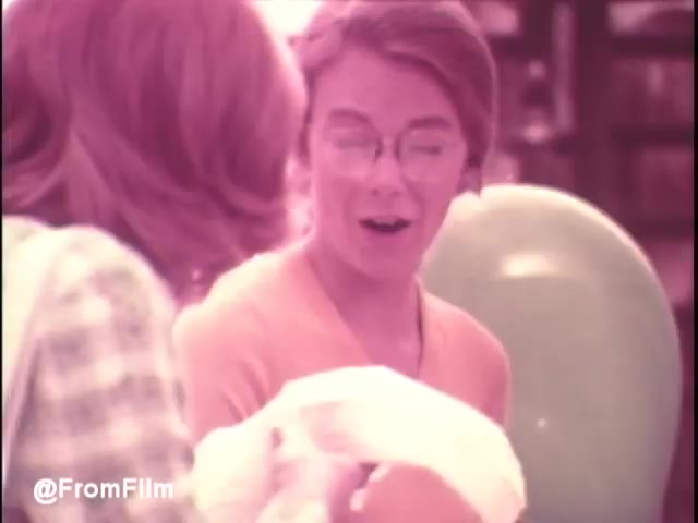 The still from the early 1970s Pamper's Diapers commercial features two people in a candid moment. One person, likely a parent, has a look of surprise or delight as they hold a diaper, while the other, possibly a friend or family member, appears engaged in conversation. The presence of a green balloon in the background adds a playful touch, characteristic of the cheerful atmosphere often found in commercials targeted at families. The color palette has a vintage feel, typical of the era's filmmaking, emphasizing warmth and nostalgia.