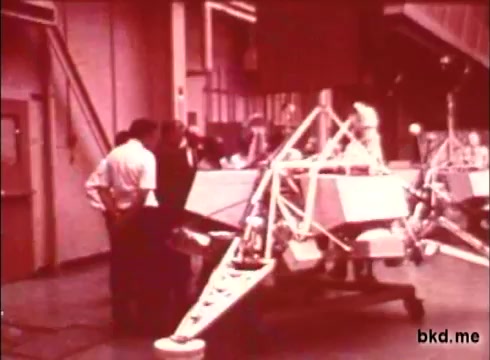 The still from the 'Apollo 12 pre-launch unknown film' shows a group of individuals, likely engineers or technicians, gathered around a lunar module prototype. The setting appears to be a hangar or assembly area, with the module prominently displayed and various equipment in the background. The scene captures a moment of collaboration and preparation ahead of the Apollo 12 mission, reflecting the attention to detail and teamwork involved in space exploration. The 16mm print likely provides a vintage look at the era's technology and the people behind these historic missions.
