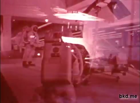 The still depicts a scene from a 16mm film related to the Apollo 12 pre-launch period. It features two astronauts in spacesuits, likely preparing for the mission. The image has a reddish tint, giving it an almost vintage or stylized appearance. In the foreground, there's a piece of equipment, possibly related to the mission, alongside visible background elements that suggest a controlled, technical environment. This film may serve an informational purpose, capturing the atmosphere and preparations leading up to the launch.