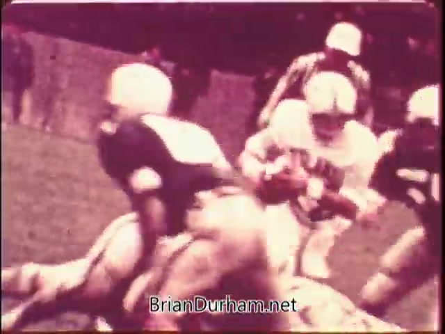 The still from the 'US Mint PSA for Silver Bicentennial coins' features a football scene, capturing a dynamic moment on the field. Players in helmets and uniforms are engaged in a tackle, highlighting the intensity of the game. The colors appear slightly faded, typical of 16mm film, contributing to a vintage aesthetic. The image suggests an energetic atmosphere, possibly connecting American culture and sports to the theme of the Bicentennial coins.