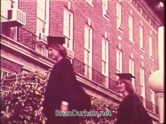 The still features two women dressed in graduation caps and gowns, walking in front of a brick building. The scene captures a celebratory moment, likely at a graduation ceremony, with emphasis on their achievements. The setting has a nostalgic tone, enhanced by the film's vintage quality.