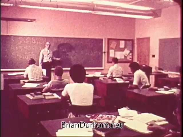 The still shows a classroom setting from a public service announcement for the US Mint's Silver Bicentennial coins. A teacher, wearing glasses, stands at the front by a blackboard, addressing a group of students seated at desks. The classroom appears retro, likely from the 1970s, with students attentively listening and taking notes.
