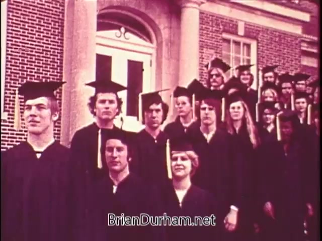 The still depicts a group of students in graduation caps and gowns, standing in front of a brick building. They appear to be celebrating a significant milestone, possibly a graduation ceremony. The setting is formal, conveying a sense of accomplishment. The students’ expressions range from serious to cheerful.