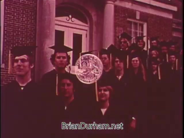 The still features a group of graduates wearing caps and gowns, posed in front of a building. They are holding a large depiction of a silver Bicentennial coin, symbolizing the commemorative coins issued by the US Mint. The scene emphasizes a celebratory atmosphere, likely highlighting the coins' significance in American history and culture.