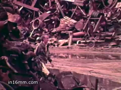 The 16mm still from 'Pat Paulsen PSA for the Environment Protection Agency' depicts a man walking through a waste site filled with discarded materials. The background features a chaotic assortment of scrap metal and refuse, highlighting the environmental impact of solid waste. The voiceover provides stark statistics about annual waste production in the U.S., emphasizing the scale of the problem and the urgent need for environmental awareness and action.