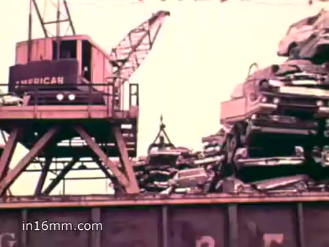The still from the 'Pat Paulsen PSA for the Environment Protection Agency' depicts a scene featuring a large crane operating over a pile of crushed cars. The crane is positioned on a platform labeled 'AMERICAN,' suggesting a focus on industrial waste and recycling. This visual emphasizes the growing problem of solid waste in the U.S., aligning with the voiceover statistics on discarded materials and the environmental impact of excess consumption. The overall tone reflects a call for awareness and action regarding waste management in the 1970s.
