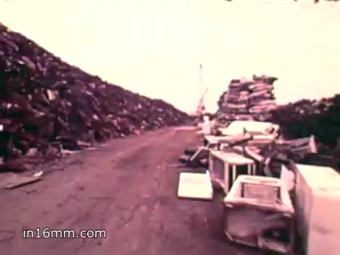 The still from 'Pat Paulsen PSA for the Environment Protection Agency' depicts a stark landscape filled with towering piles of solid waste, including a variety of discarded items like appliances and vehicles. The scene emphasizes the overwhelming scale of waste generated annually in the U.S., aligning with the voiceover's statistics on the massive quantities of cans, bottles, plastics, and other materials contributing to environmental concerns during the 1970s. The road through the waste conveys a sense of neglect and urgency regarding the need for environmental protection.