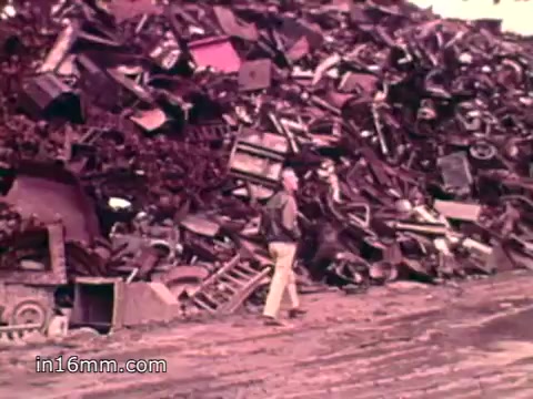 The still depicts a large, chaotic pile of discarded items, including metal scraps and various consumer goods, highlighting the overwhelming amount of waste generated in the U.S. during the 1970s. In the foreground, a person walks along a dirt path, seemingly dwarfed by the sheer scale of the waste. This visual serves as a stark reminder of the environmental challenges posed by consumerism, aligning with the voiceover's statistics on waste production and its impact.