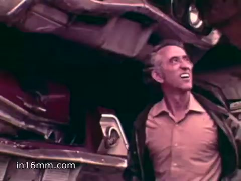 The still features a middle-aged man smiling and looking upward in a scrapyard filled with discarded cars. The scene emphasizes the issue of solid waste, highlighting the extensive amount of trash produced in the U.S. and the environmental concerns associated with it, as conveyed by the voiceover statistics regarding waste disposal.