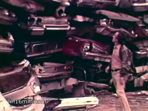 The still features a man standing amidst a towering pile of junked cars in a junkyard, symbolizing waste accumulation. This visual aligns with the voiceover statistics about the significant waste produced annually in the U.S., highlighting environmental concerns related to solid waste. The scene communicates a message about the impact of consumerism and the pressing need for environmental awareness and action in the 1970s.