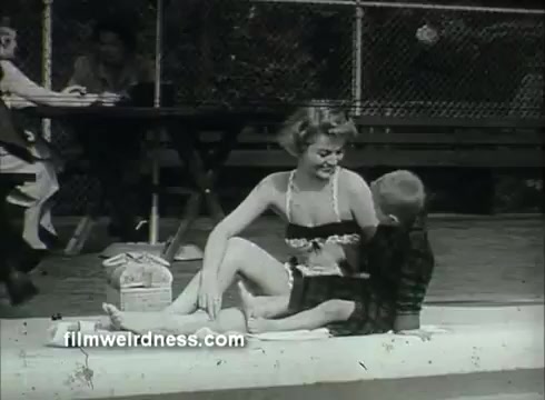 The still from the US Air Force PSA features a woman in a swimsuit sitting by a pool, engaging with a young boy. She appears to be smiling and interacting warmly with him. In the background, there are people seated at a picnic table, emphasizing a social setting typical of the 1950s-1960s era. The overall scene evokes a sense of leisure and family-friendly summer activities.