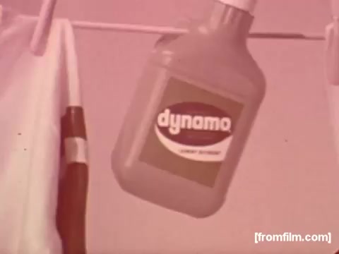 The still from the 'Colgate Dynamo Liquid Detergent Commercial' features a bottle of Dynamo liquid detergent prominently displayed. The label is clear, showcasing the brand name 'Dynamo' in bold letters. The color scheme is simple, likely reflecting the design trends of the 1970s. The background appears soft and warm-toned, which adds a nostalgic feel to the advertisement. Overall, the image effectively communicates the product's brand identity in a straightforward manner.