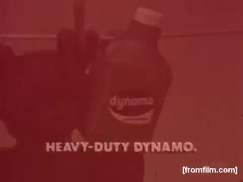 The still features a close-up of a bottle of Colgate Dynamo liquid detergent, prominently displaying the label. The background is likely to be blurred, drawing focus to the product. The text 'HEAVY-DUTY DYNAMO' is displayed in bold, emphasizing the detergent's strength and effectiveness. The overall aesthetic reflects the style of 1970s commercials, likely using warm tones to appeal to viewers.