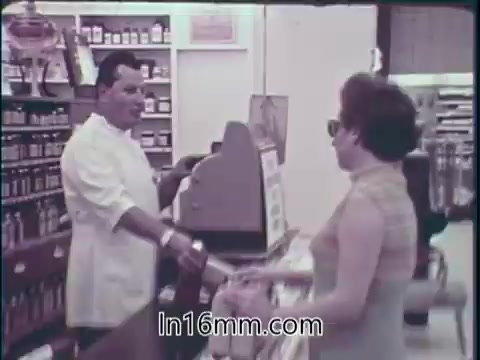 The still from the 1960s PSA features a man in a white coat behind a store counter, interacting with a woman who is looking up at him while holding a shopping bag. The background displays shelves lined with various jars and products, highlighting a vibrant marketplace atmosphere. The image conveys a sense of trust and community in the American retail experience, emphasizing the availability of safe food and effective drugs. The overall tone aligns with the era's optimistic view of consumerism and health.