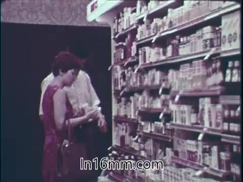 The still from the 1960s PSA features a woman in a purple dress examining products on a supermarket shelf, filled with various food items and medications. The advertisement promotes the abundance and safety of consumer goods in the American marketplace, highlighting the effectiveness of drugs for health and beauty. It marks the 30th anniversary of the Food and Drug Administration, emphasizing the agency's role in ensuring product quality for consumers.