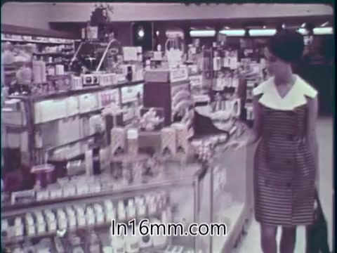 The still from the 1960s PSA highlights a grocery or drugstore aisle filled with a variety of products, including food and drugs. A woman in a striped dress with a white collar browses the display, emphasizing consumerism in the American marketplace. The imagery reflects the era's focus on the abundance of available goods, promoting the notion of safety and effectiveness in both food and pharmaceuticals. The tone suggests optimism about the benefits these products bring to everyday life.
