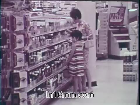 The still from the 1960s PSA features a woman and a child in a grocery store aisle, surrounded by various consumer products. The shelves are stocked with food items and over-the-counter drugs. The woman, wearing a colorful patterned outfit, appears to be examining products while the child, in a striped dress, is looking at the shelves below. The scene conveys a sense of abundance in the American marketplace, emphasizing the availability of both food and health-related products during that era.