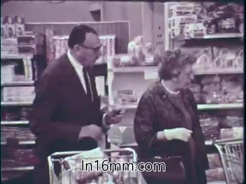 The still features two individuals, a man and a woman, shopping in a grocery store filled with various products on the shelves. The man, dressed in a suit with glasses, appears to be discussing something with the woman, who is carrying a handbag. Their shopping carts are visible, suggesting they are engaged in selecting goods. This scene reflects a mid-20th century American marketplace, emphasizing the wide availability of food and medicine, which aligns with the public service announcement's themes of consumer safety and health awareness. The overall atmosphere conveys a sense of trust in the products available to consumers.