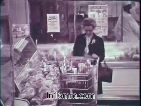 The still from the 1960s FDA PSA depicts a woman shopping in a grocery store. She is standing beside a display of various food items, filling her shopping cart. The environment suggests a focus on the abundance of safe and effective products available in the American marketplace. The overall tone promotes the idea of consumer confidence in food and drugs, celebrating the anniversary of FDA regulations.