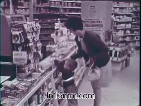 The still from the 1960s PSA by the Food and Drug Administration shows a woman shopping in a grocery store with a young child. The shelves are stocked with various food products and medicines, emphasizing the abundance and variety available in the American marketplace. The message highlights the safety and effectiveness of food and drugs, portraying them as beneficial for health and beauty. The scene reflects a time of optimism about consumer products and their role in American life.