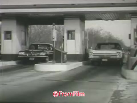 The still depicts a scene from a 1960s Aetna public safety message about speeding and dangerous driving. It shows two cars—one entering and one exiting a toll plaza. The setting reflects the period's automotive culture, emphasizing themes of road safety and responsible driving. The black-and-white film adds a nostalgic tone, aligning with the safety message's serious nature.