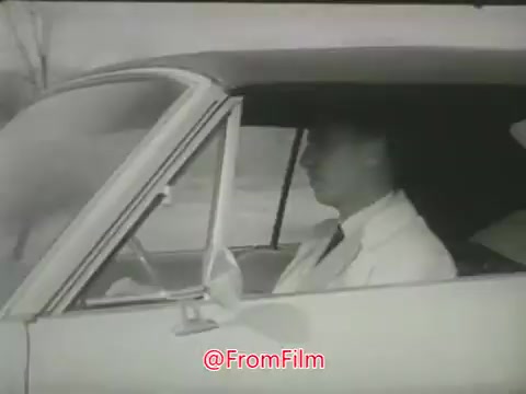 The still depicts a man sitting in the driver's seat of a car, wearing a light-colored suit. He appears focused on the road ahead. The image reflects the tone and style of the 1960s, showcasing the concerns about speeding and dangerous driving through a somber, serious atmosphere typical of public safety messages from that era. The black-and-white format enhances the vintage feel of the clip.