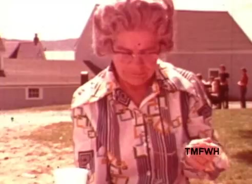The still from 'Birdseye New England Style Vegetables' features an older woman outdoors, focused on a small item in her hand. She has gray hair styled in soft curls and wears glasses, accompanied by a colorful patterned shirt. In the background, there are hints of structures, possibly a barn or farmhouse, and people can be seen at a distance, suggesting a communal or family gathering in a rural setting. The overall vibe reflects a nostalgic and wholesome atmosphere.