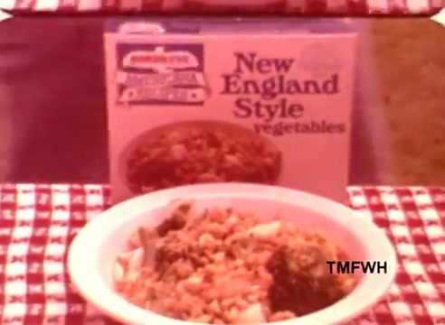 The still features a bowl of cooked vegetables prominently displayed in the foreground, showcasing a mix that likely includes broccoli and other assorted vegetables. Behind it is a box of Birdseye New England Style Vegetables, highlighting the product branding. The background shows a checkered tablecloth, enhancing the homey, rustic aesthetic typical of mid-20th century food advertisements. The overall composition suggests a focus on convenience and wholesome eating.