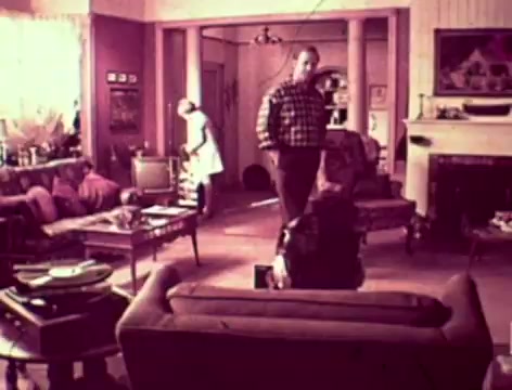 In this 16mm still from a 1970s Listerine commercial, a father expresses concern about his son’s silence, questioning whether he has bad breath or is out of touch. The mother suggests trying Listerine, explaining its benefits beyond just treating colds. The scene captures a typical family living room of the era, with the father standing while the mother is engaged in household activity, reflecting a relatable dynamic of parental concern.
