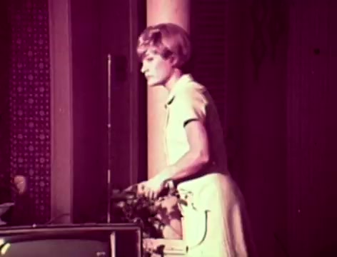 The still from the 1970s Listerine commercial shows a woman in a light-colored dress, standing near a television. The background features patterned wallpaper and a subtle domestic setting. The scene captures a moment of concern as a father questions his son's silence, while the mother suggests using Listerine to address his bad breath. The visual aesthetic aligns with the era, utilizing warm tones and a vintage feel.