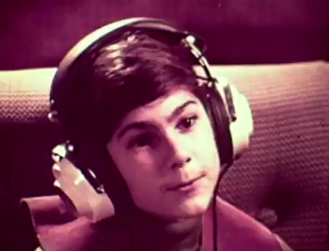 The still from the Listerine commercial features a young boy wearing large headphones, seated on a couch. He appears contemplative, possibly listening to a conversation or music. The context suggests a family dynamic where the father worries about communication with his son, hinting at issues like bad breath as a potential barrier. The mother introduces Listerine as a solution, emphasizing its effectiveness against germs that cause bad breath. The retro aesthetics and setting reflect the 1970s style of advertising.
