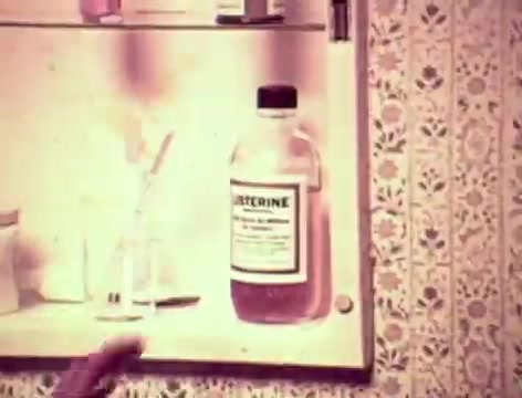 The still depicts a bathroom cabinet with a bottle of Listerine prominently displayed. The background features vintage wallpaper with floral patterns. A hand reaches towards the Listerine, suggesting the mother’s encouragement for the father to use it. The scene captures a typical family dynamic, highlighting concerns about communication and personal hygiene, with a focus on the product’s effectiveness against bad breath.