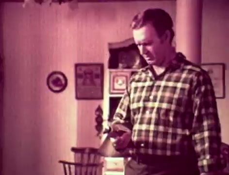 The still features a man in a plaid shirt, looking pensive as he holds a bottle, presumably of Listerine. He stands in a domestic setting that includes a simple wooden chair and framed pictures on the wall, suggesting a homey atmosphere. The dialogue captures a moment of concern from a father about communication with his son, reflecting themes of family interaction and personal hygiene in a lighthearted manner. The overall tone is nostalgic, characteristic of 1970s advertising.