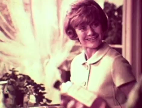 The still from the 1970s Listerine commercial features a mother smiling at her husband as she suggests using Listerine to combat bad breath. The setting appears warm and domestic, with soft lighting and a background of plants and curtains, emphasizing a comforting family environment. The mother's demeanor is cheerful and encouraging, while the father's expression indicates concern about his relationship with their son and his self-image. The dialogue highlights themes of communication and personal hygiene.