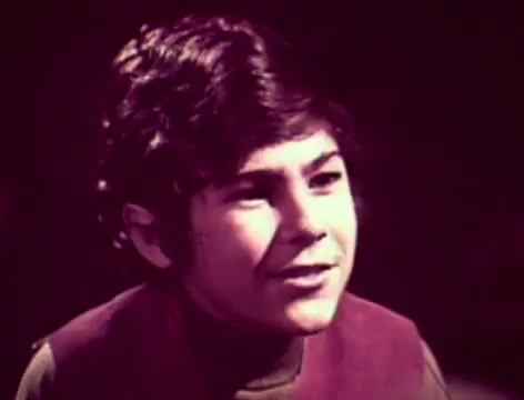 The still is from a 1970s Listerine commercial featuring a young boy speaking to his father. The boy has a thoughtful expression, indicating he may be considering what was just said. The scene conveys a sense of concern about personal communication and hygiene, with the father wondering about his relationship with his son and possibly his breath. The dialogue hints at the importance of using Listerine to address bad breath, alongside a touch of humor about its strength. The visuals likely have a vintage aesthetic typical of commercials from that era.