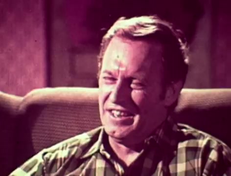 In this 16mm still from a 1970s Listerine commercial, a father, dressed in a plaid shirt, is seated on a couch with a concerned expression. The context of the scene revolves around his worries about why his son hasn't spoken to him in weeks, leading to concerns about bad breath and being perceived as 'square.' The mother suggests Listerine, emphasizing its dual purpose of treating colds and combating bad breath, prompting a humorous acknowledgment from the father about its strong taste. The overall tone captures a relatable family dynamic while highlighting the product's benefits.