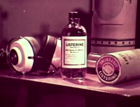 The 16mm still from the 1970s Listerine commercial features a vintage setup with a glass bottle of Listerine prominently displayed, alongside an old-fashioned container of Listerine and a pair of black and white headphones. The color palette is muted, characteristic of the era, and the overall atmosphere conveys a domestic scene. The dialogue suggests a familial concern about communication and hygiene, with a focus on the strong effects of Listerine in combating bad breath.