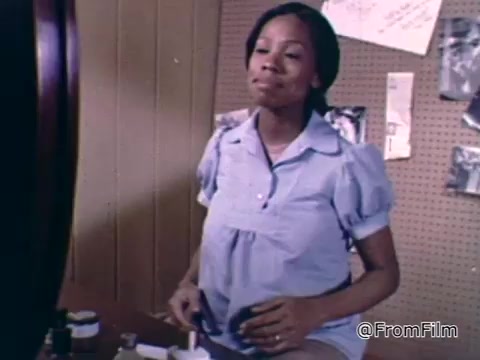 The still features a pregnant woman smiling as she looks into a mirror. She is wearing a light blue, short-sleeved blouse, and appears to be engaged in a personal grooming routine. The background suggests a domestic setting with various items and notes pinned to a bulletin board. The image conveys a sense of care and positivity, likely emphasizing themes of maternal health and wellness relevant to the March of Dimes campaign.