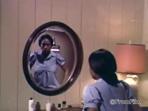 The still from the 1975 PSA by the National Foundation of the March of Dimes features a woman looking into a round mirror in a simple interior setting. She has a contemplative expression, and the scene is marked by a warm atmosphere created by soft lighting from a nearby lamp. The focus on her reflection suggests themes of self-reflection and awareness, likely connected to the PSA's message about health or maternal care.