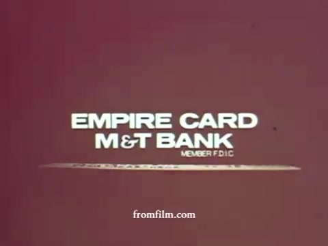 The still features a simple and bold graphic design with the text 'EMPIRE CARD' prominently displayed above 'M&T BANK,' emphasizing the bank's branding. The background is a muted shade, contributing to a retro 1970s aesthetic. It also includes 'MEMBER FDIC' to highlight the bank's affiliation and credibility. The overall composition is straightforward and focused on promoting the Empire Card service.