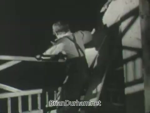 The still depicts a scene with a figure in overalls, possibly a child, on a wooden structure or staircase. The setting is dimly lit, suggesting a sense of suspense or action. The figure appears to be engaged in some activity related to climbing or exploring the structure. Overall, the image captures a moment full of potential adventure or drama typical of promotional material from the era.