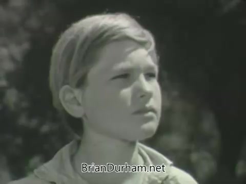 The still features a young boy with short, light-colored hair, looking thoughtfully into the distance. His expression suggests contemplation or concern. The backdrop appears natural, possibly indicating an outdoor setting. This moment captures the essence of childhood reflection often portrayed in television promos from that era.