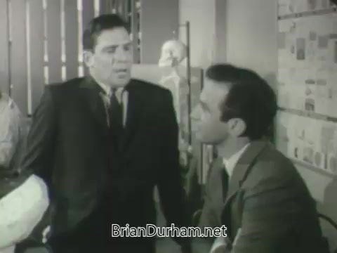 The still features two men in suits engaged in a conversation. One man appears to be standing and animated, while the other is sitting and listening intently. The background includes elements indicative of an office or conference setting, with various posters or charts visible. The overall tone suggests a professional or dramatic context, typical of a promotional segment for a television network's upcoming season.